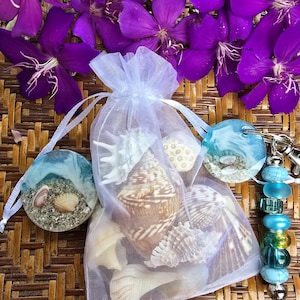 May include: A collection of seashells in a white mesh bag, with two round resin keychains featuring beach scenes. The keychains have blue and white resin, with sand and shells. Purple flowers are in the background.