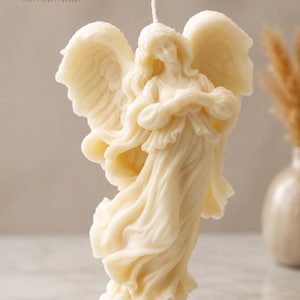 Könnte beinhalten: Eine cremefarbene Wachskerze in Form eines Engels mit großen, detaillierten Flügeln und fließenden Gewändern. Die Figur steht auf einer wolkenartigen Basis mit einem einzelnen weißen Docht am Kopf. Das Logo ELVIDA CANDLES ist deutlich zu sehen.