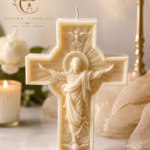 Könnte beinhalten: Eine cremefarbene Wachskerze in Kreuzform mit einem Relief einer Figur mit ausgebreiteten Armen, einer Taube darüber und floralen Details am Fuß. Die Kerze hat einen einzelnen Docht an der Oberseite und zeigt den Text ELVIDA CANDLES.