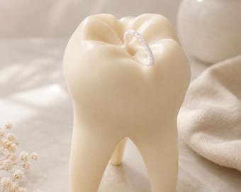 May include: An off-white candle shaped like a human molar tooth with a single white wick centered on the top surface. The candle features a smooth, matte finish and stands on two distinct roots. The logo ELVIDA CANDLES LIGHT SCENT MOMENT is visible.