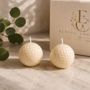Könnte beinhalten: Zwei cremefarbene, kugelförmige Kerzen mit einer strukturierten, genoppten Oberfläche, die an einen Golfball erinnert, jeweils mit einem weißen Docht. Daneben steht eine weiße Pappschachtel mit dem goldenen Logo ELVIDA CANDLES und dem Text LIGHT SCENT MOMENT.