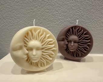 May include: Two round candles with celestial designs. One is ivory, featuring a sun and moon face. The other is brown, with a similar design. Both candles have a wick at the top, and are on a white surface.