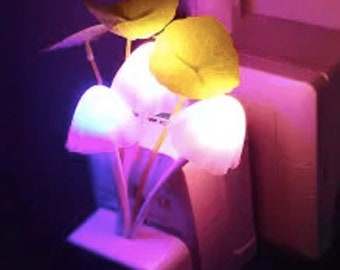 May include: A decorative night light shaped like a cluster of glowing mushrooms and leaves. The light emits a soft, multi-colored glow, with shades of pink, blue, and yellow. The night light plugs directly into a standard wall outlet.
