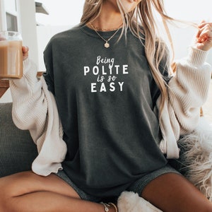 Może przedstawiać: Ciemnoszara koszulka z białym napisem "Being Polite is so Easy". Koszulka ma okrągły dekolt. Osoba nosi kremowy kardigan i szorty. Trzyma szklankę jasnego napoju.