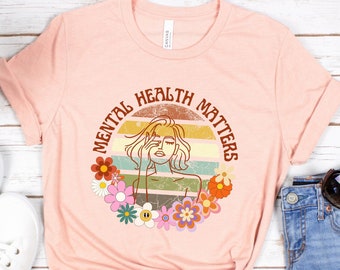May include: A pink t-shirt with a retro-style graphic design featuring a woman with flowers and the text "Mental Health Matters".