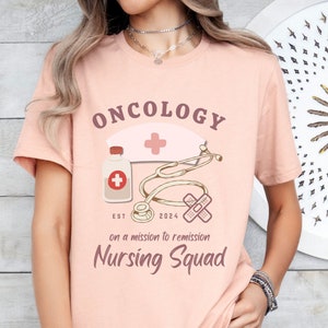 May include: Pink t-shirt with the text "Oncology Nursing Squad" and a graphic of a nurse's hat, a stethoscope, a bottle of pills, and a bandage. The text "Est 2024" and "On a mission to remission" are also included.