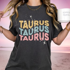 May include: A dark grey t-shirt with a retro design featuring the word "Taurus" repeated three times in different colors and a constellation graphic. The shirt is being worn by a person with long blonde hair.
