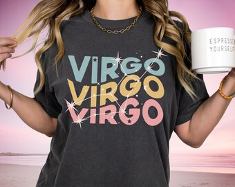 Może przedstawiać: Szary t-shirt z napisem "Virgo" powtórzonym trzy razy w kolorowym, warstwowym tekście. Każde "Virgo" jest obwiedzione na biało i otoczone konstelacją gwiazd. Modelka trzyma biały kubek z napisem "Espresso Yourself".