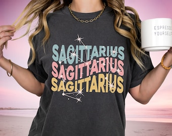 Könnte beinhalten: Ein dunkelgraues T-Shirt mit dem Wort "Sagittarius" dreimal in verschiedenen Farben wiederholt. Das Wort ist in einer Retro-Schriftart mit Sternen und einem Sternbild-Design.