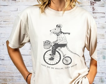 Pode incluir: Uma camiseta bege com um gráfico de um rato andando de bicicleta com uma cesta de flores. O texto na camiseta diz "They see me rollin', they hatin'".