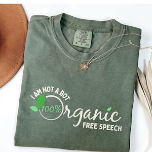 May include: An olive green t-shirt with the text "I AM NOT A BOT 100% Organic FREE SPEECH" in white. A silver necklace with a circular pendant rests on the shirt. A brown hat and dried plant are in the background.