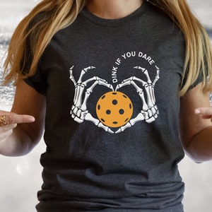 May include: A dark gray t-shirt with a white skeleton hand design holding a yellow pickleball. The text "Dink if you dare" is written in a circle around the pickleball.