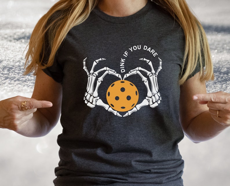 May include: A dark gray t-shirt with a white skeleton hand design holding a yellow pickleball. The text "Dink if you dare" is written in a circle around the pickleball.