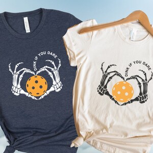 May include: Two t-shirts with a skeleton hand design holding a pickleball. The text on the shirts reads "Dink if you dare".