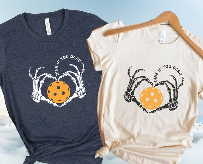 May include: Two t-shirts with a skeleton hand design holding a pickleball. The text on the shirts reads "Dink if you dare".