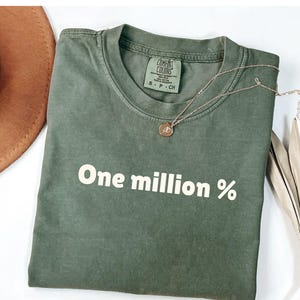 以下が含まれることがあります： 前面に白い文字で「One million %」がプリントされた緑色のTシャツです。