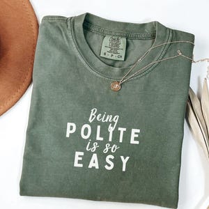 May include: A green t-shirt with the text "Being Polite is so Easy" in white lettering. The shirt is folded and laid on a white surface.