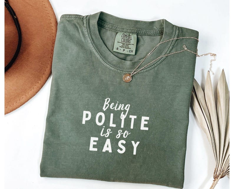 May include: A green t-shirt with the text "Being Polite is so Easy" in white lettering. The shirt is folded and laid on a white surface.