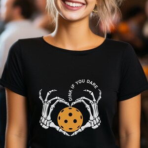 May include: Black t-shirt with a white graphic of two skeleton hands holding a pickleball in a heart shape. The text "Dink if you dare" is written in a circle around the hands.