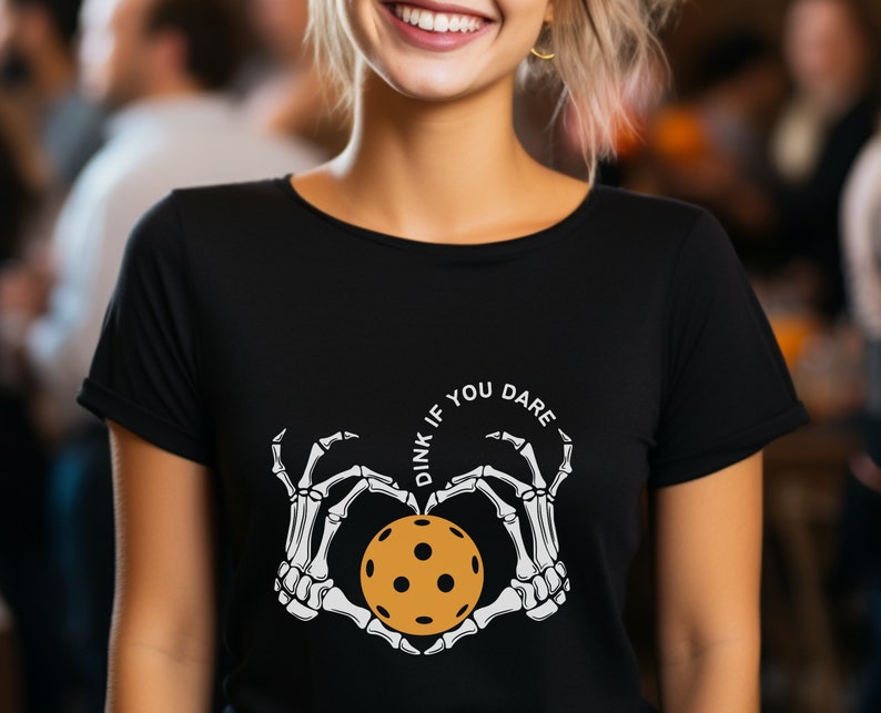 May include: Black t-shirt with a white graphic of two skeleton hands holding a pickleball in a heart shape. The text "Dink if you dare" is written in a circle around the hands.