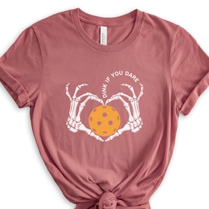 May include: A pink t-shirt with a white skeleton hand design holding a yellow pickleball. The text "Dink if you dare" is written in a circle around the pickleball.