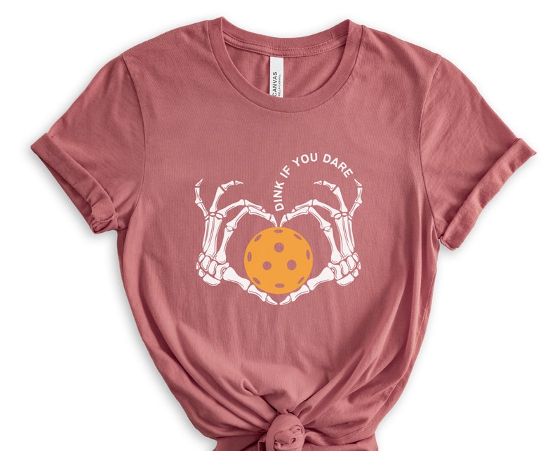 May include: A pink t-shirt with a white skeleton hand design holding a yellow pickleball. The text "Dink if you dare" is written in a circle around the pickleball.