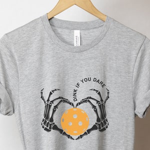 May include: Gray t-shirt with a black skeleton hand design holding a yellow pickleball. The text "Dink if you dare" is written in a circle around the pickleball.