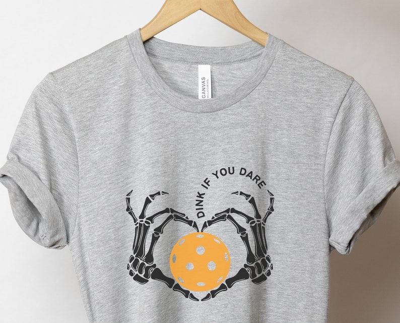 May include: Gray t-shirt with a black skeleton hand design holding a yellow pickleball. The text "Dink if you dare" is written in a circle around the pickleball.