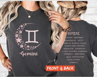 May include: A dark gray t-shirt with a pink floral design and the Gemini zodiac symbol. The back of the shirt has the word "Gemini" and a list of personality traits associated with the zodiac sign.