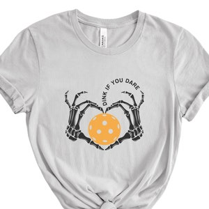 May include: A light gray t-shirt with a black skeleton hand design holding a yellow pickleball. The text "Dink if you dare" is written in a circle around the pickleball.