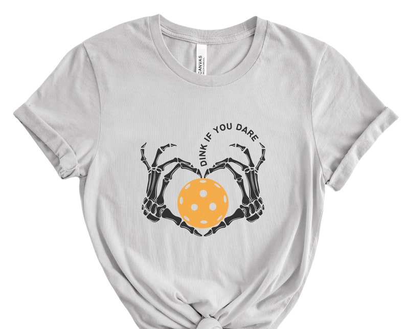 May include: A light gray t-shirt with a black skeleton hand design holding a yellow pickleball. The text "Dink if you dare" is written in a circle around the pickleball.