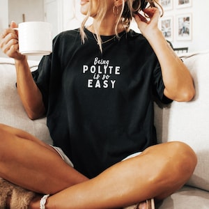 May include: A woman wearing a black t-shirt with the text "Being Polite Is So Easy" in white lettering. She is sitting on a couch and holding a white mug.