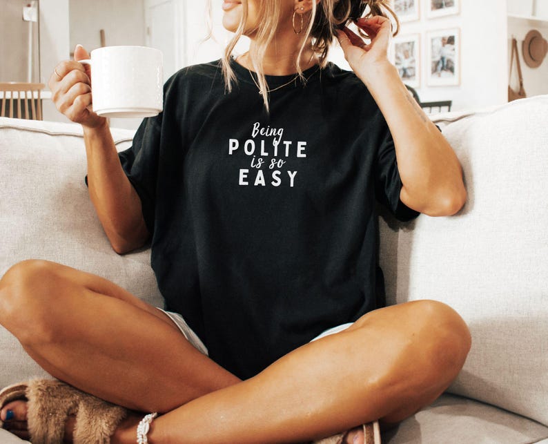 May include: A woman wearing a black t-shirt with the text "Being Polite Is So Easy" in white lettering. She is sitting on a couch and holding a white mug.
