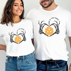 May include: Two white t-shirts with a skeleton hand design holding a yellow pickleball. The text on the shirts reads "Dink if you dare".