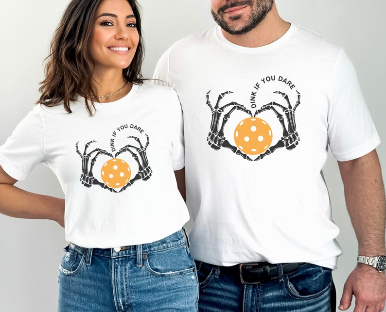 May include: Two white t-shirts with a skeleton hand design holding a yellow pickleball. The text on the shirts reads "Dink if you dare".