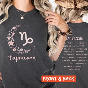 May include: A dark grey t-shirt featuring a pink floral design and the zodiac symbol for Capricorn. The back of the shirt displays the word "Capricorn" and a list of personality traits associated with the zodiac sign.