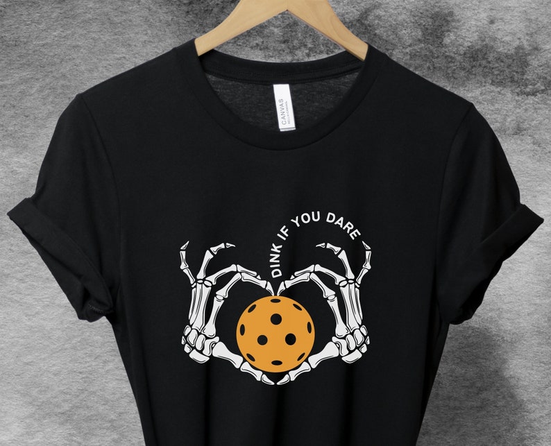 May include: Black t-shirt with a white skeleton hand design holding a yellow pickleball. The text "Dink if you dare" is written in a circle around the pickleball.