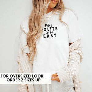 May include: White t-shirt with the text "Being Polite is so Easy" in black lettering. The model is wearing the shirt with a cardigan and grey pants. The text "For Oversized Look - Order 2 Sizes Up" is at the bottom of the image.