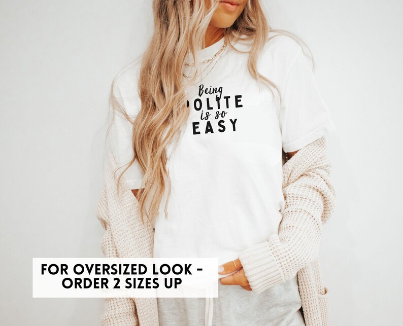 May include: White t-shirt with the text "Being Polite is so Easy" in black lettering. The model is wearing the shirt with a cardigan and grey pants. The text "For Oversized Look - Order 2 Sizes Up" is at the bottom of the image.
