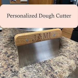 Puede incluir: Un cortador de masa personalizado con un mango de madera grabado con el nombre "JAMI". La cuchilla de acero inoxidable es rectangular. El texto "Personalized Dough Cutter" está en un rectángulo rosa claro.