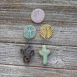 May include: Five ceramic ornaments, two are round with geometric designs in shades of blue, yellow, and pink, and two are crosses in shades of blue, green, and brown. The ornaments are displayed on a rustic wooden background.