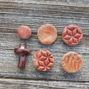 May include: Six ceramic tiles with different designs. The tiles are in shades of pink and brown. The designs include a cross, a geometric pattern, and a leaf pattern.