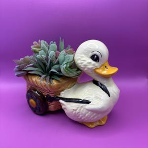 May include: A white ceramic duck planter with a yellow beak and black eyes, pulling a brown wicker wagon with a green succulent plant inside.