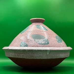 May include: A pink ceramic tagine with a light blue leaf pattern. The tagine has a wide, round base and a domed lid.