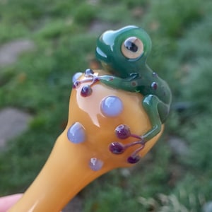 May include: A hand-blown glass pipe with a frog perched on a mushroom-shaped bowl. The pipe has a yellow stem and bowl, decorated with purple and blue accents. The frog is green with dark spots and large eyes.