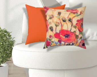 May include: Two decorative throw pillows on a white chair. One pillow is solid orange, the other features a floral design with red poppies, green stems, and yellow and blue accents against a cream background. The pillows add a pop of color to the room.