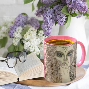 May include: A pink-handled coffee mug with a portrait of a woman with blue eyes and curly hair. The mug is filled with a dark liquid, and sits on a wooden tray next to an open book and eyeglasses. Lilac flowers are in the background.