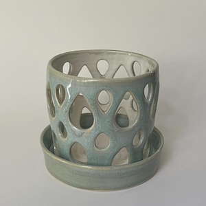 Puede incluir: Una maceta de cerámica azul claro con un diseño de corte decorativo. La maceta tiene un platillo a juego.