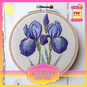 May include: A hand embroidery pattern featuring two purple irises with green leaves and stems. The pattern is displayed in a wooden embroidery hoop. The text "HAND EMBROIDERY PATTERN" is displayed below the hoop.