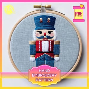 Może przedstawiać: Wzór do haftu ręcznego przedstawiający łamacza orzechów z niebieskim kapeluszem, czerwoną kurtką i białą brodą. Wzór jest wyświetlany w drewnianej obręczy do haftu na jasnoniebieskim tle z tkaniny. Tekst "HAND EMBROIDERY PATTERN" jest wyświetlany pod obręczą.